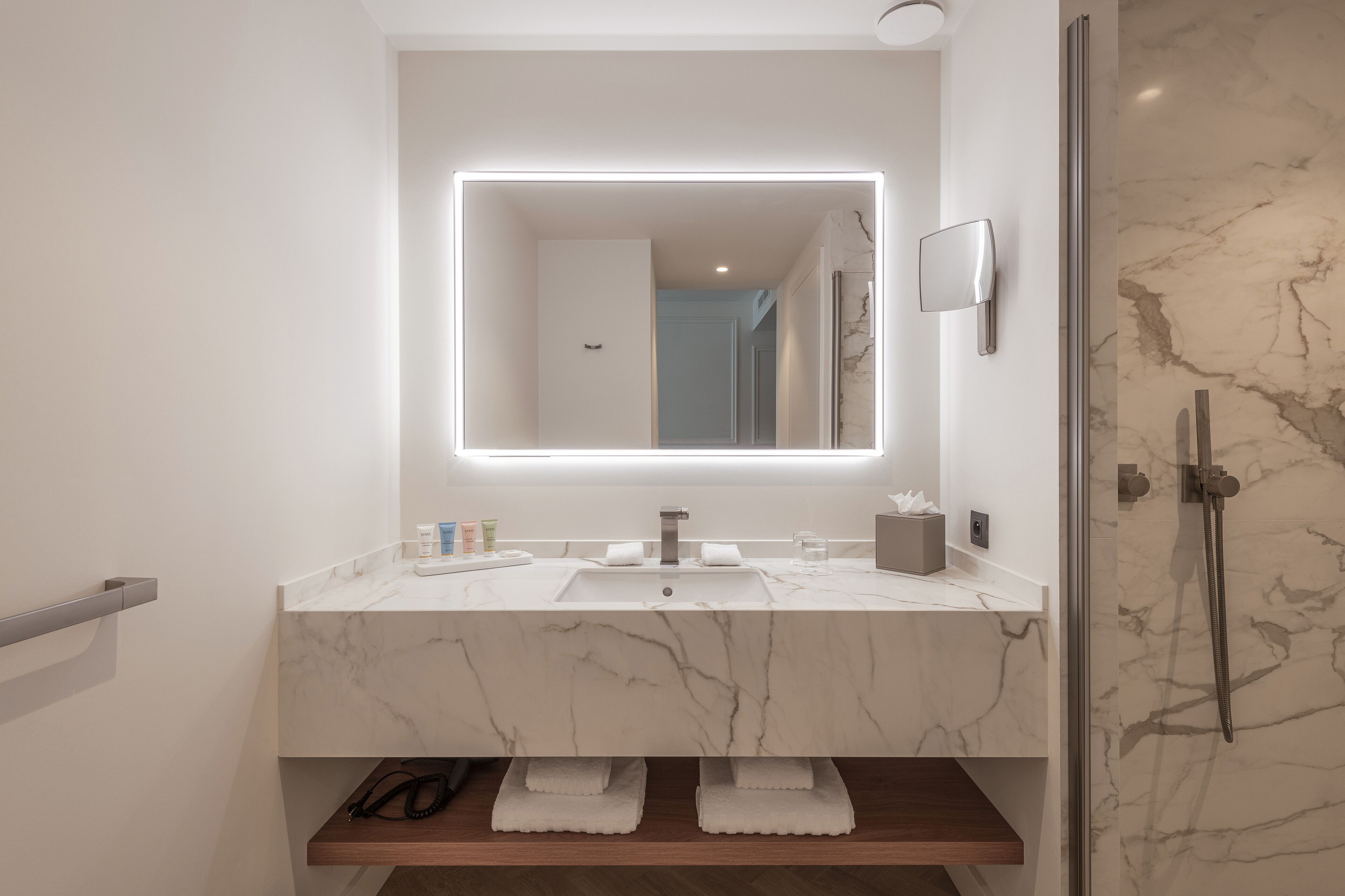 Un baño moderno con lavamanos de mármol, un espejo grande con iluminación integrada y una ducha con mampara de vidrio.
