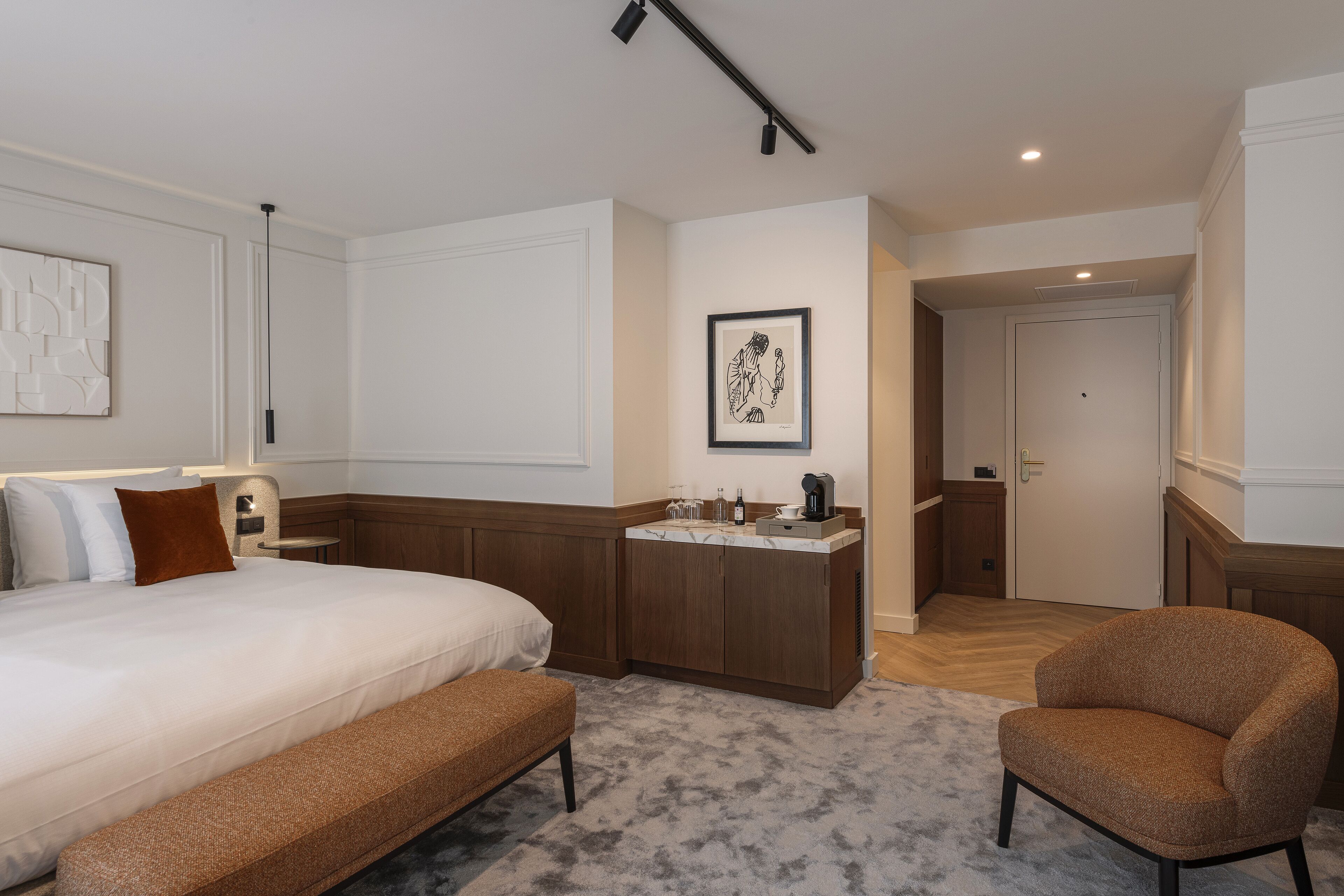 A hotel room with a large bed, a bench, a chair, a small kitchenette, and a door leading to another room.