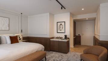 Une chambre d’hôtel avec un grand lit, un banc, une chaise, une petite kitchenette et une porte donnant sur une autre pièce.