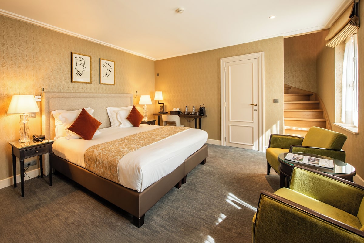 Ein Hotelzimmer mit einem großen Bett, zwei Nachttischen inklusive Lampen, einem kleinen Tisch mit einer Zeitschrift, einem grünen Sessel und einer Treppe.