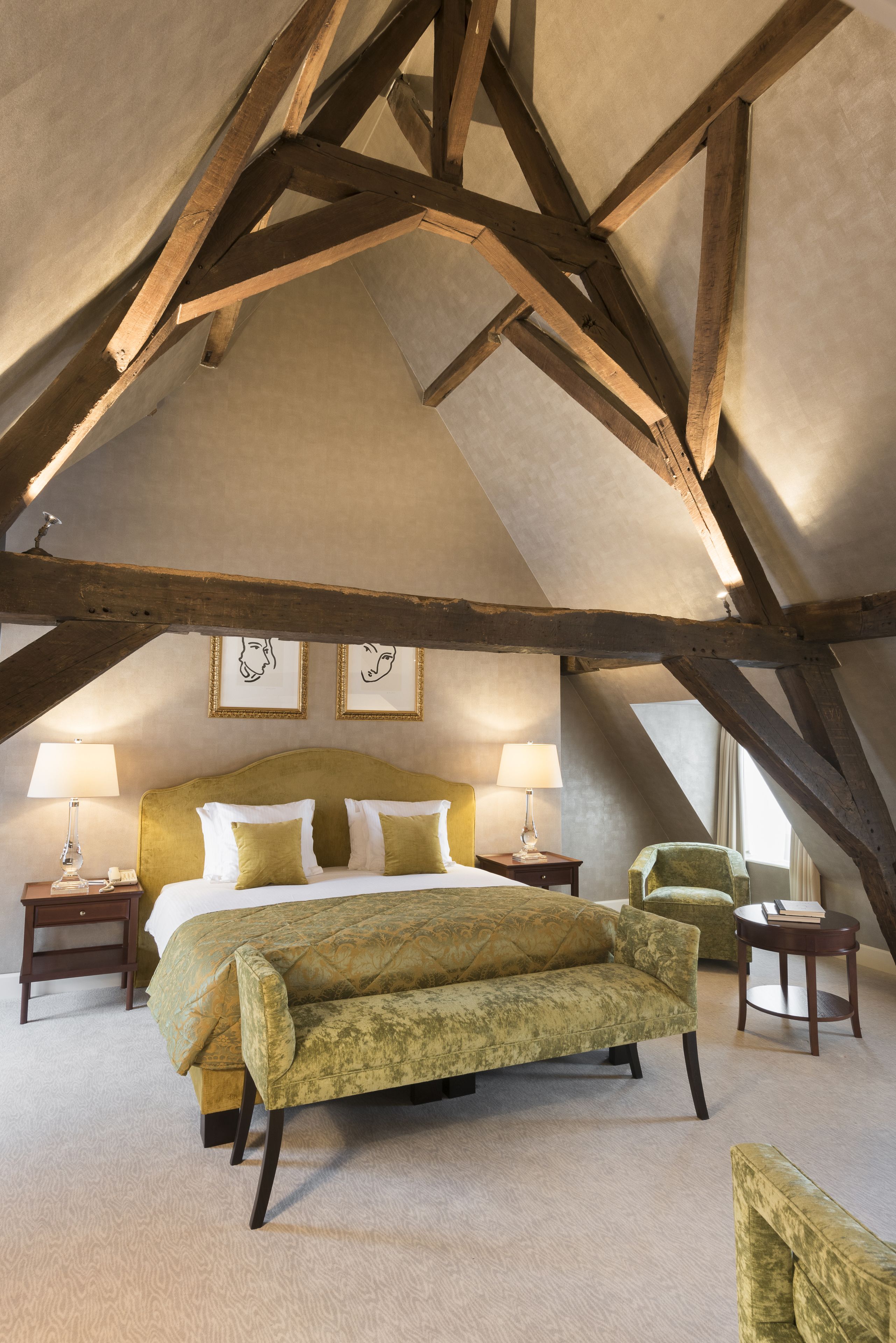 A bedroom with a high ceiling, exposed wooden beams, a large bed with green bedding, two bedside tables with lamps, and framed artwork on the wall.
