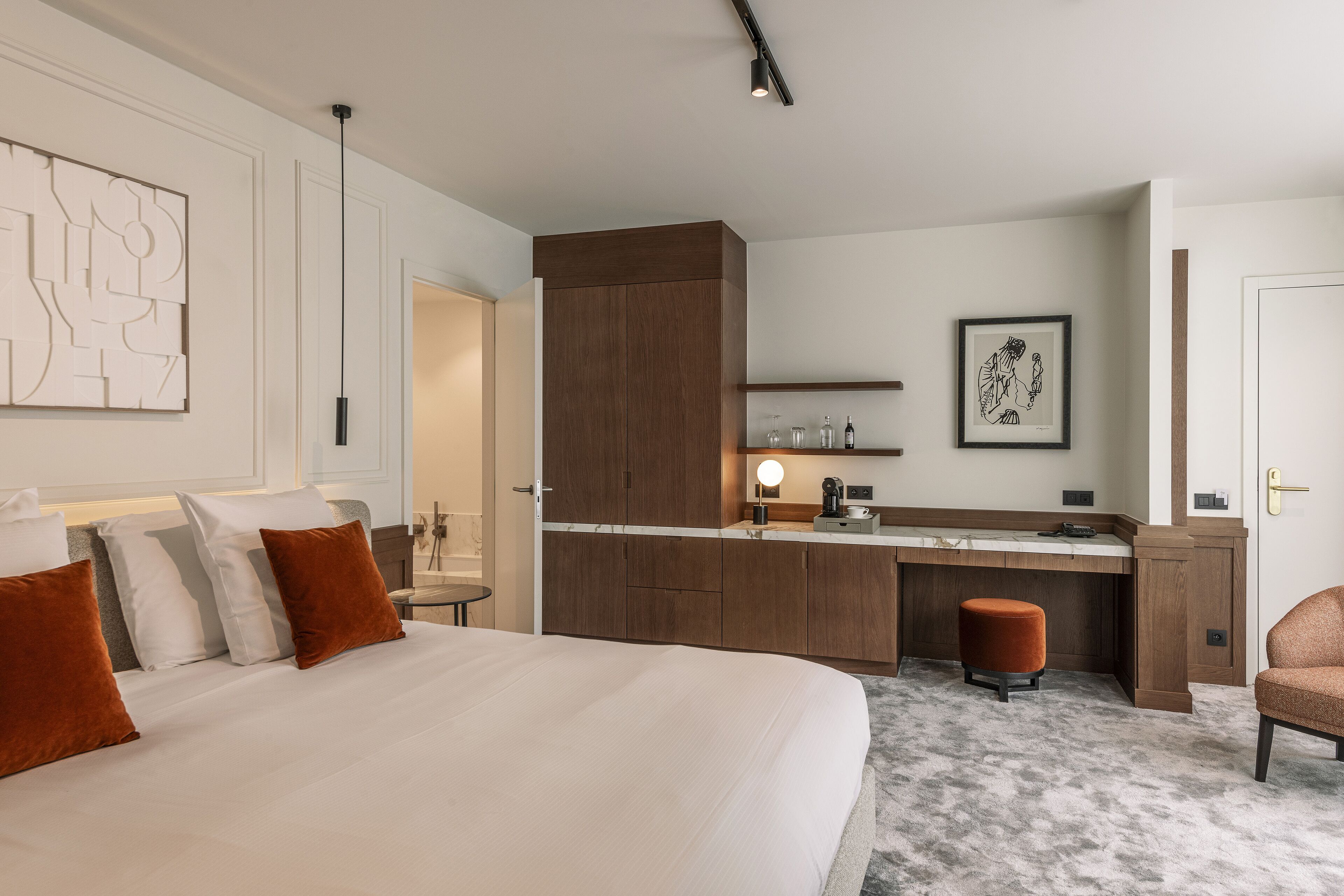 A hotel room with a large bed, a desk, and a chair. There is a small stool and a framed picture on the wall.