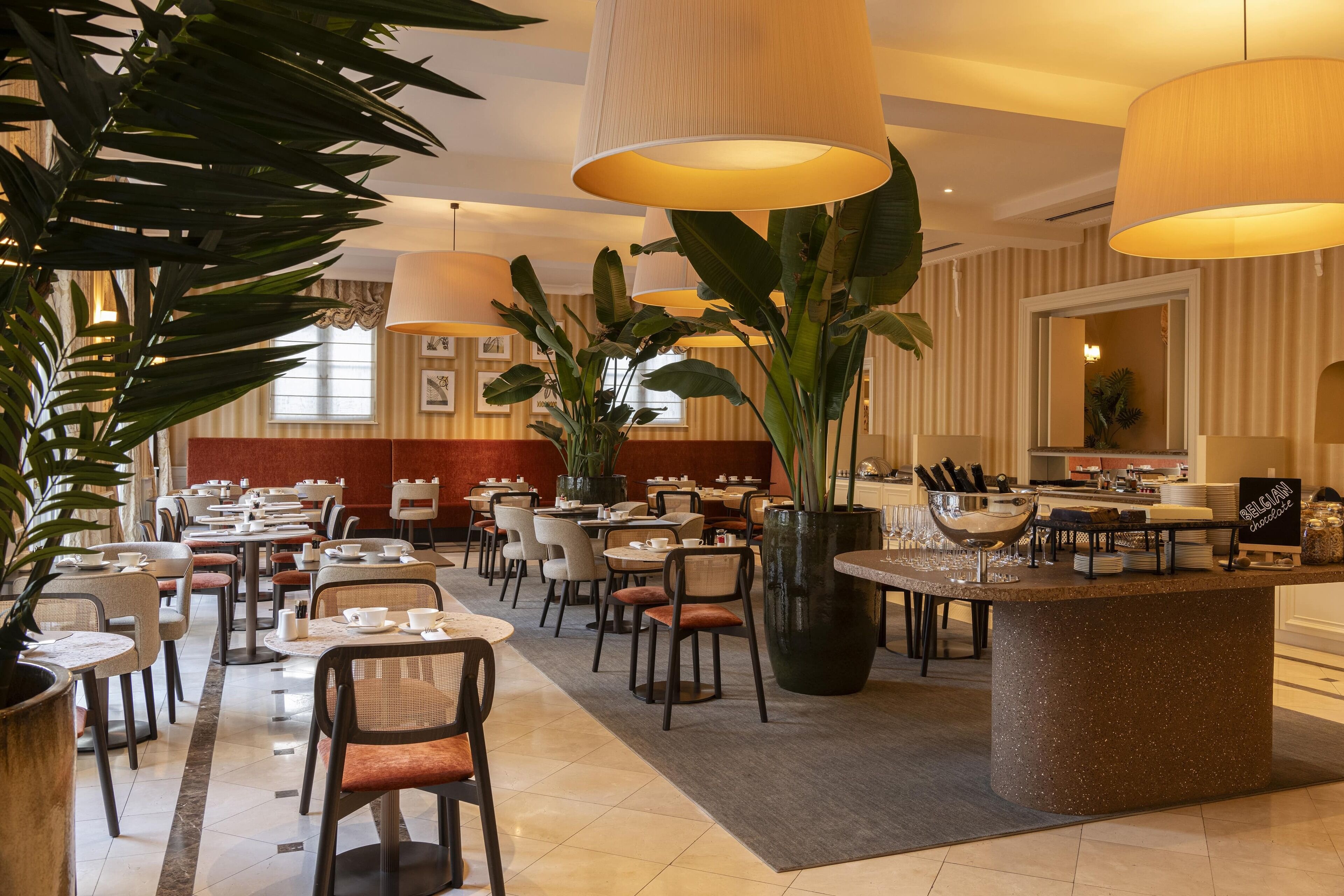 A hotel lobby with multiple dining tables, chairs, and large potted plants.