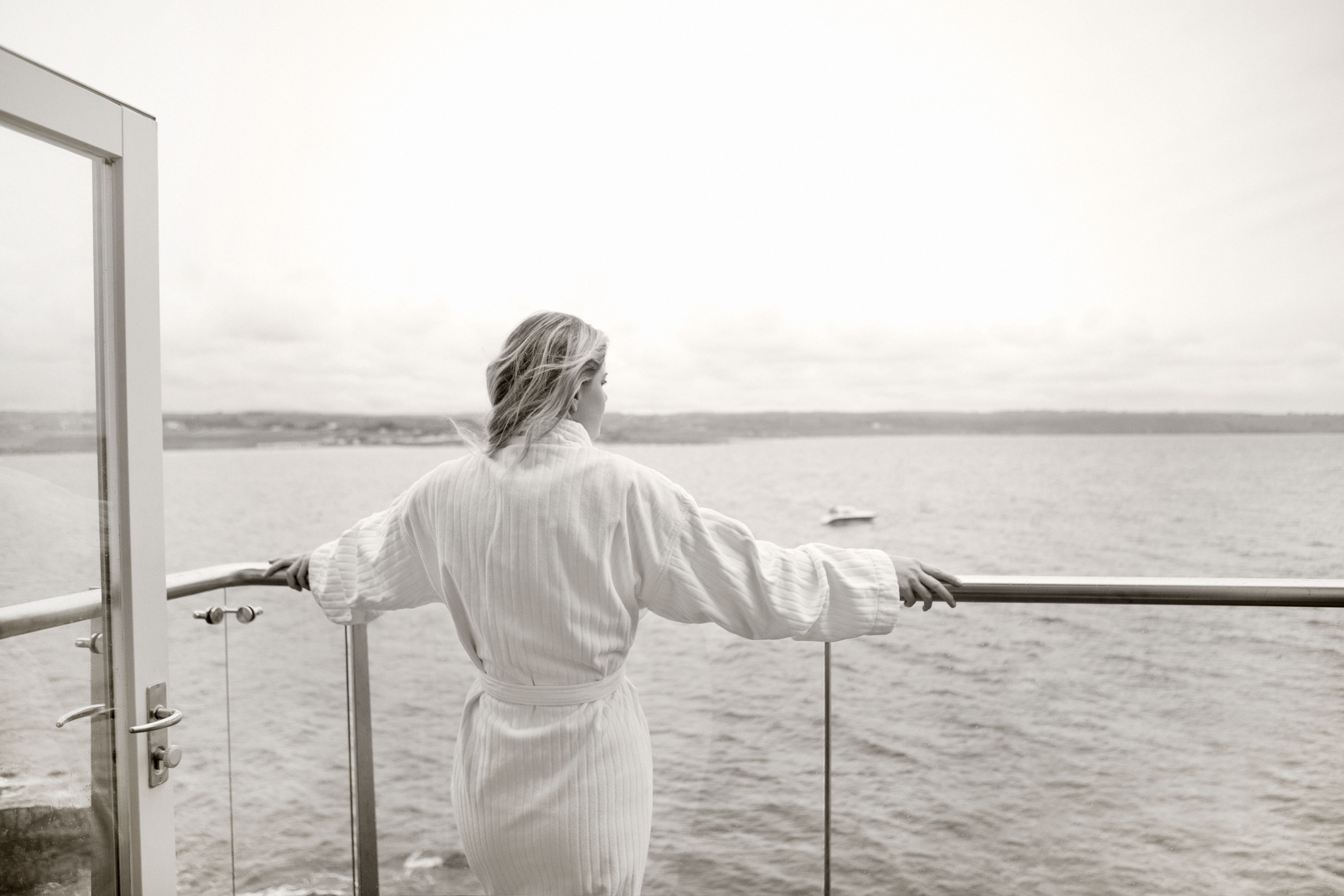 Une personne vêtue d’une longue robe blanche se tient près d’une balustrade, surplombant une étendue d’eau dont on aperçoit la rive au loin.