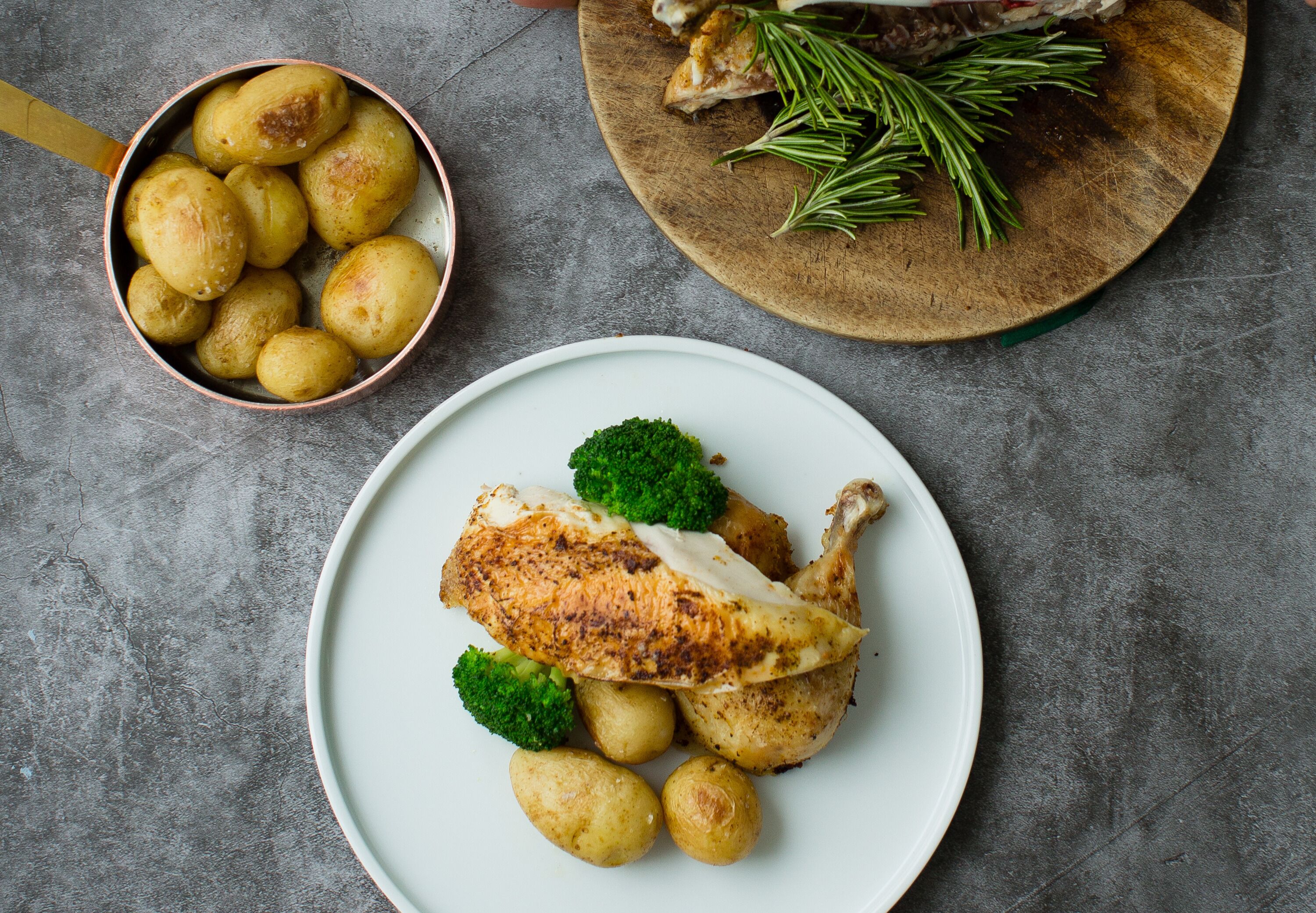 A plate with roasted chicken, broccoli, and potatoes on a gray surface.
