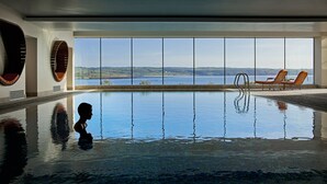 Una amplia piscina interior con vistas a un lago y colinas. Hay dos sillones de descanso junto a la piscina.