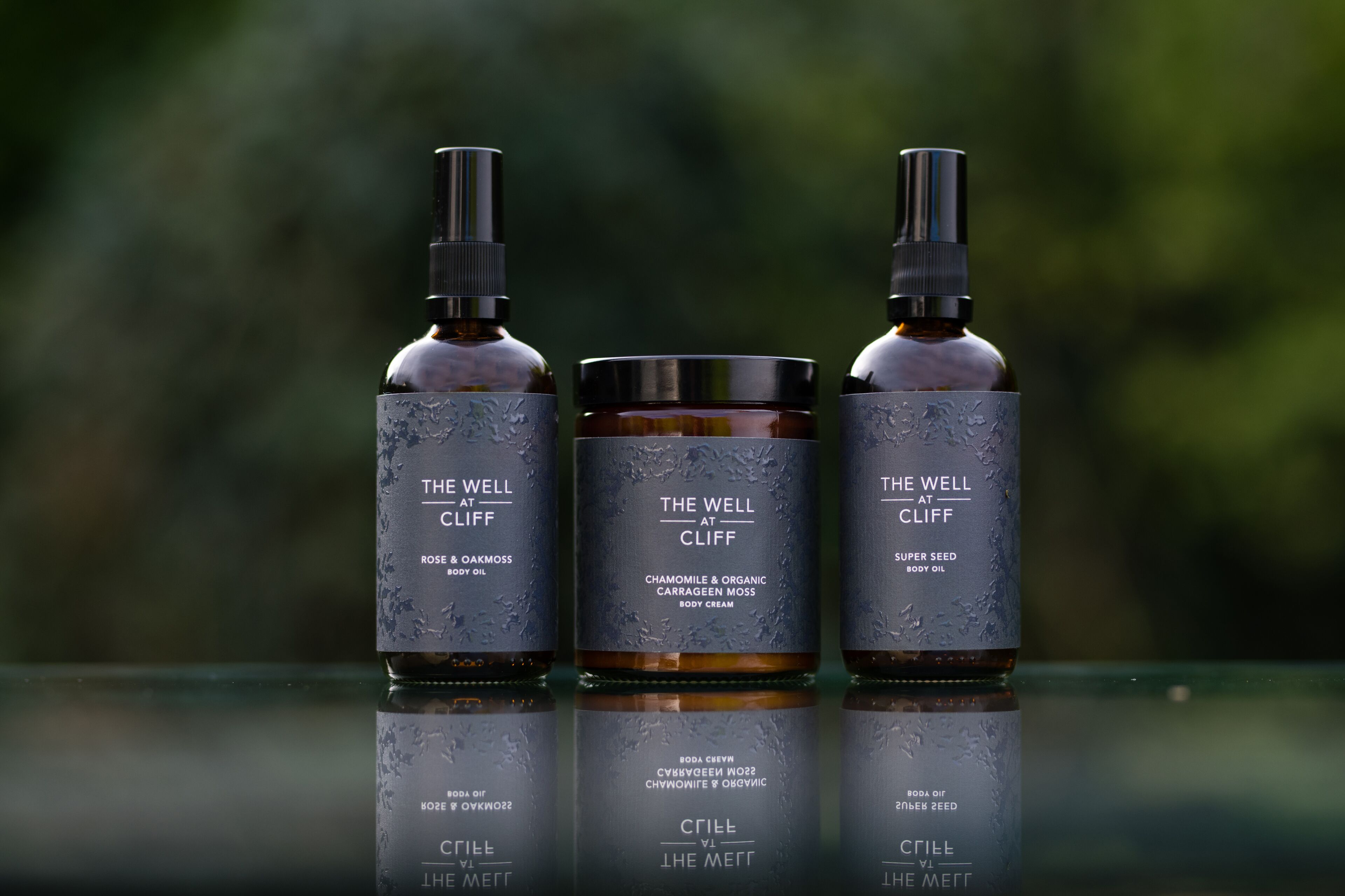 Three bottles of body oil on a reflective surface with a blurred natural background.