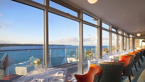 A dining area with tables set for a meal, large windows offering a view of the sea, and a balcony with a railing. - Cliff House Hotel (Ardmore)