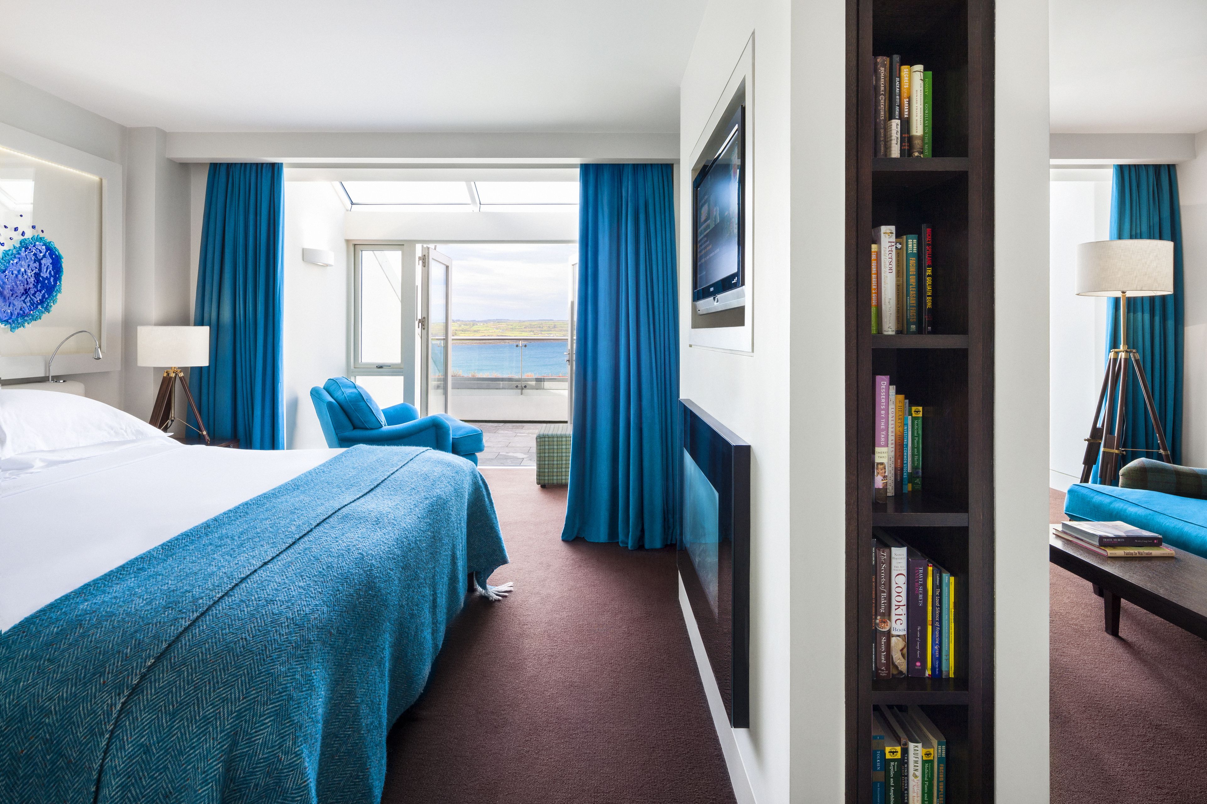 A hotel room with a large bed, a seating area with a sofa and chairs, a bookshelf, and a view of the sea through a glass door.