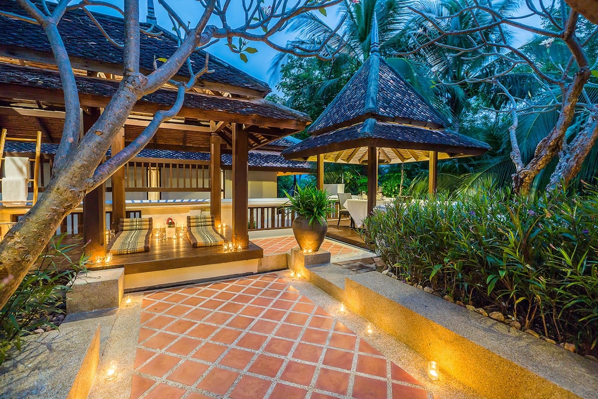 A traditional wooden structure with a tiled patio, surrounded by lush greenery and palm trees.