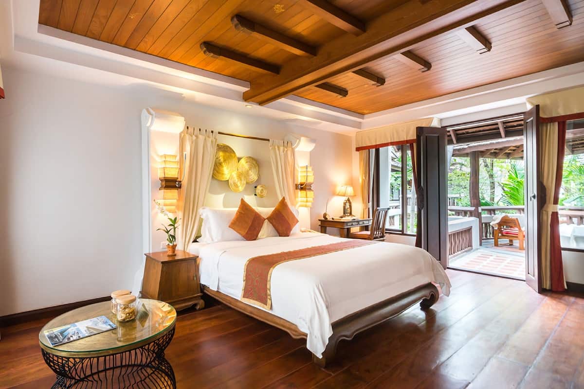A spacious bedroom with a large bed, wooden ceiling beams, a glass coffee table, and a view of the outdoors.