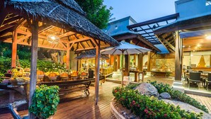 An outdoor dining area with wooden tables and chairs, a thatched-roof structure, and potted plants. - Muang Samui Spa Resort (Koh Samui)