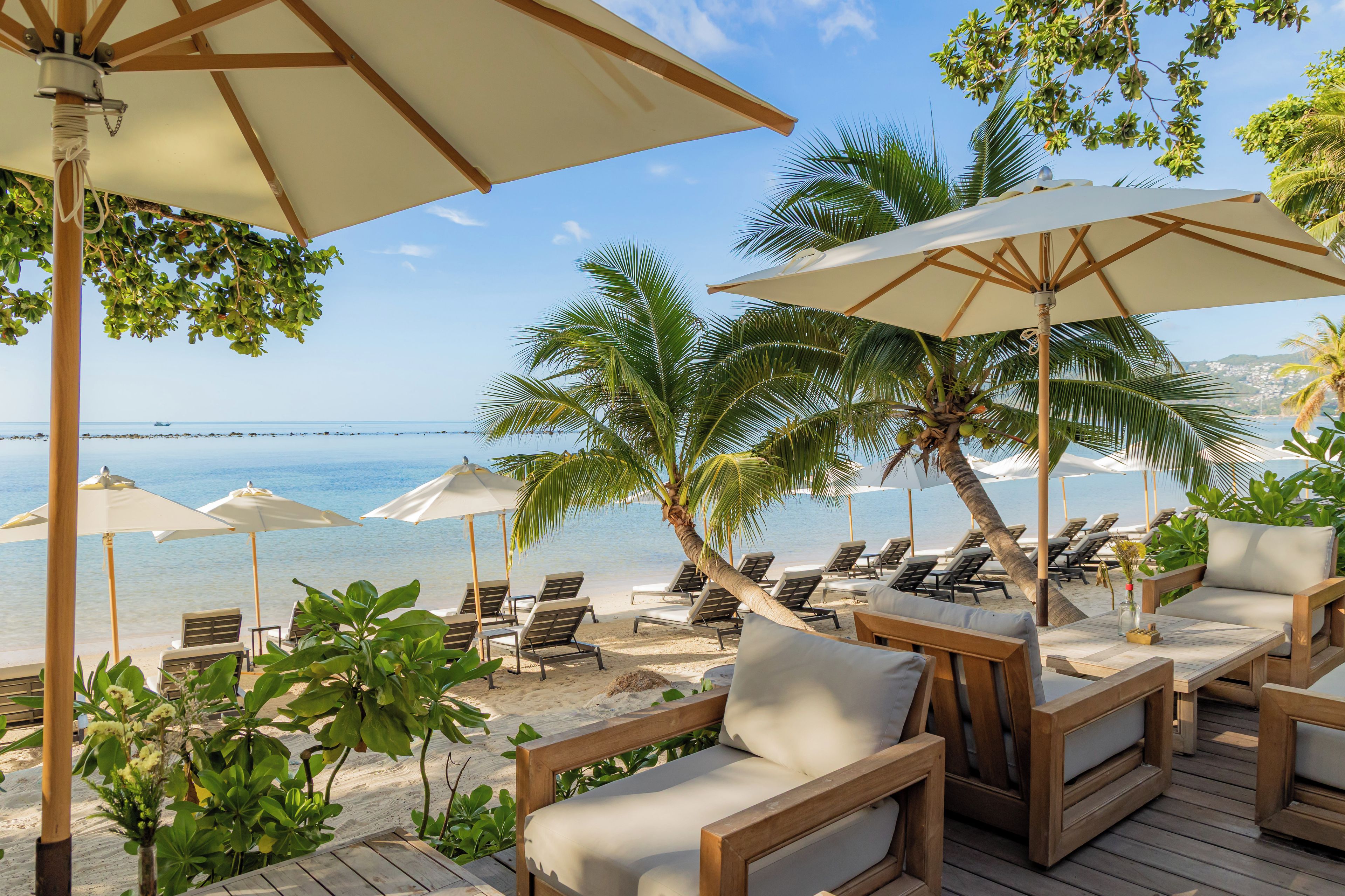 Ein Strandloungebereich mit Holzmöbeln, weißen Kissen und großen Sonnenschirmen, die Schatten spenden.