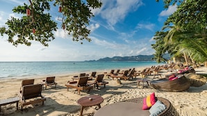A beach with wooden lounge chairs and wicker seating, a sandy shore, and a view of the ocean and distant mountains. - Muang Samui Spa Resort (Koh Samui)