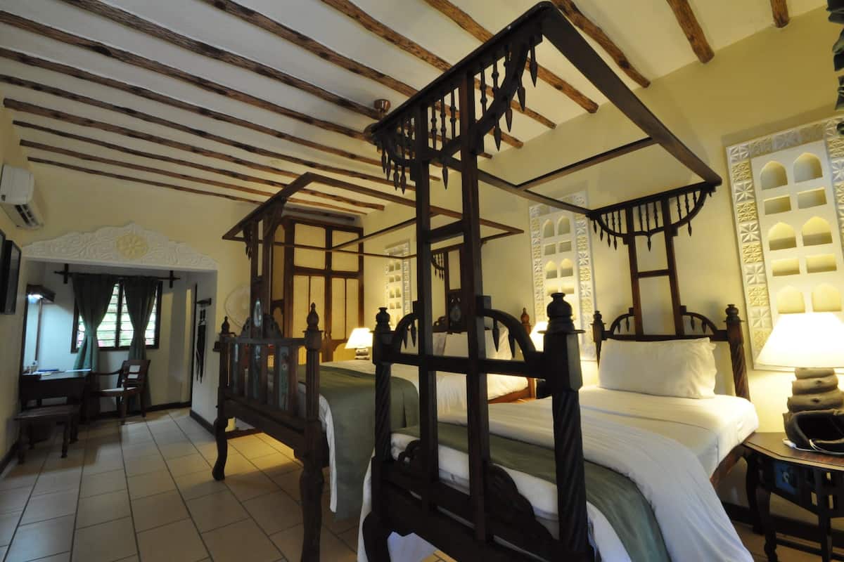 A traditional four-poster bed with a wooden frame and white bedding, a small table with a lamp, and a chair in a room with tiled flooring and wooden beams on the ceiling.