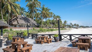Une terrasse en bois, agrémentée de tables de pique-nique et de plantes en pot, qui offre une vue sur une plage bordée de palmiers et sur un bâtiment à toit de chaume.