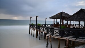 Une jetée en bois, surmontée de constructions à toits de chaume, mène à une zone de plage aménagée avec des sièges et des tables.