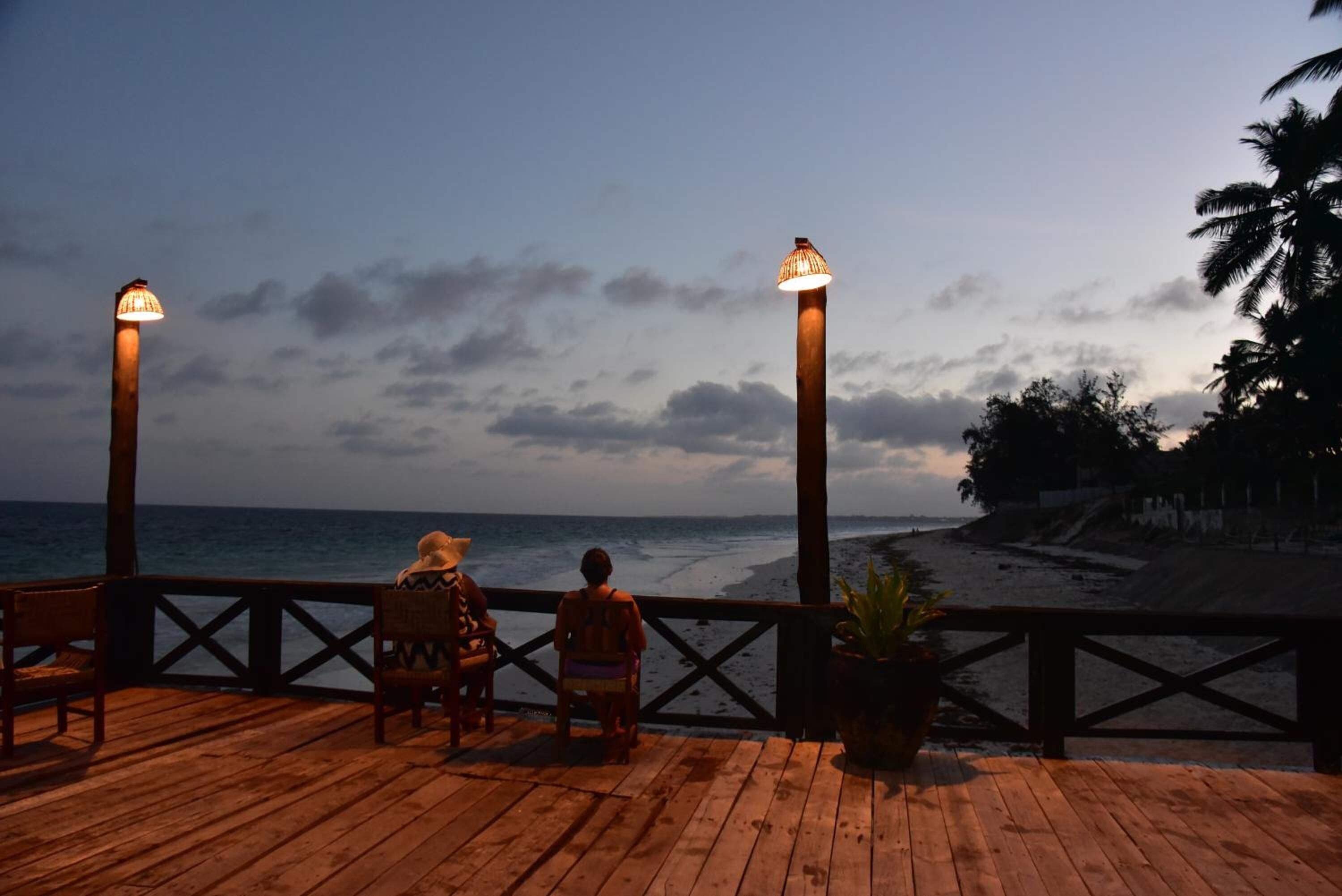 Una terraza de madera con dos personas sentadas, con vista a una playa al anochecer.