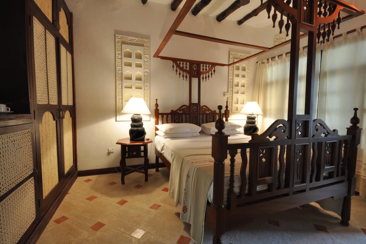 A traditional four-poster bed with a wooden frame and a canopy, two bedside tables with lamps, a decorative wall panel, and a tiled floor.