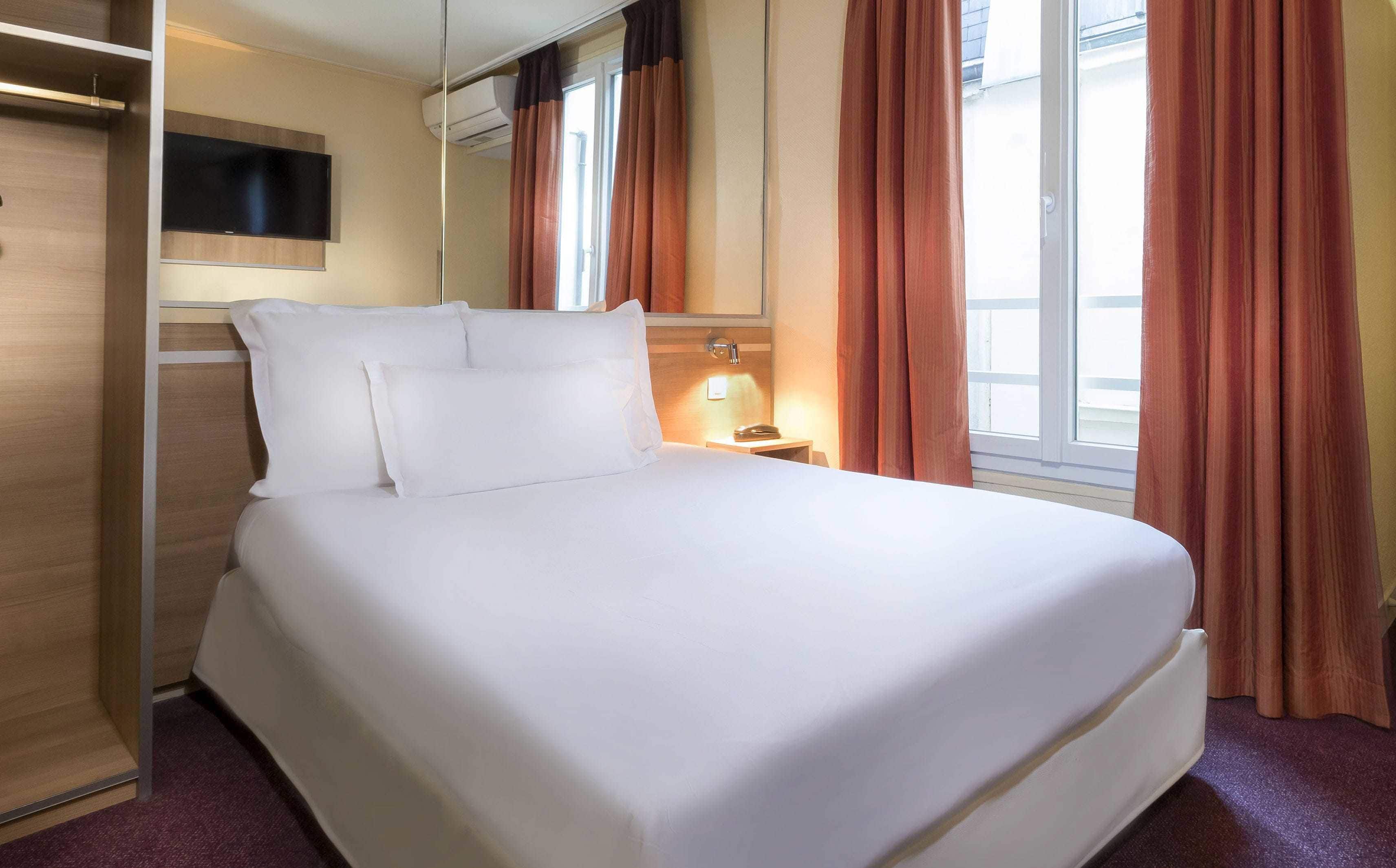 Ein Hotelzimmer mit Bett, Nachttisch mit Lampe, Wand-Fernseher und einem Fenster mit Vorhängen.