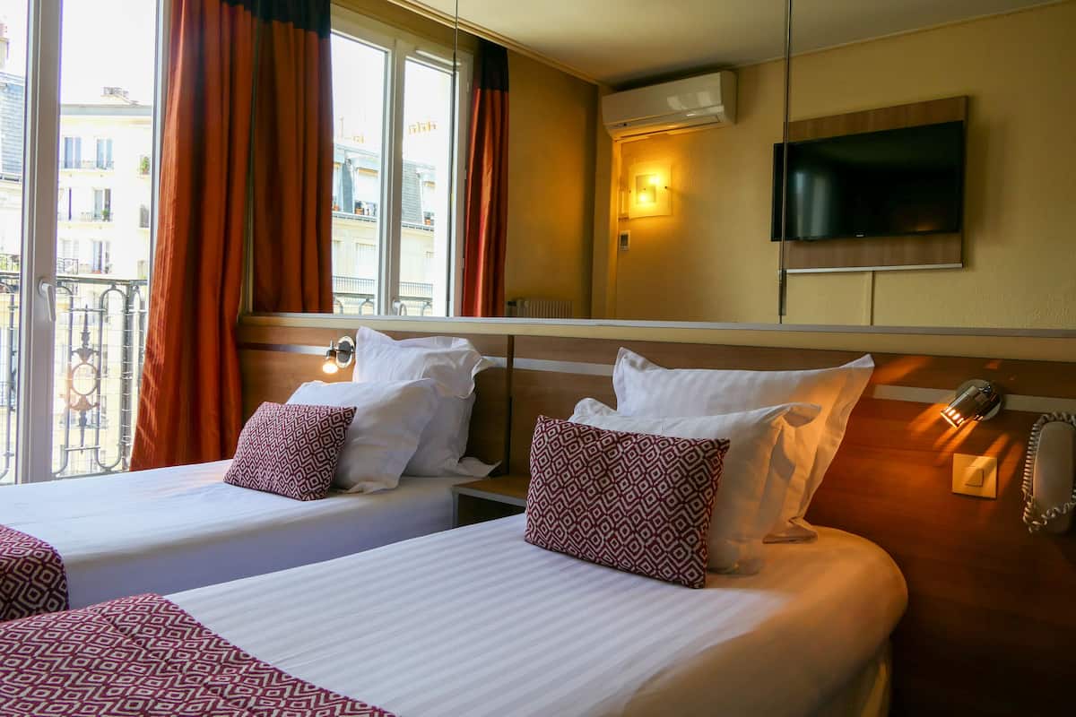Een hotelkamer met twee bedden, een aan de muur bevestigde televisie en een balkon met gordijnen.