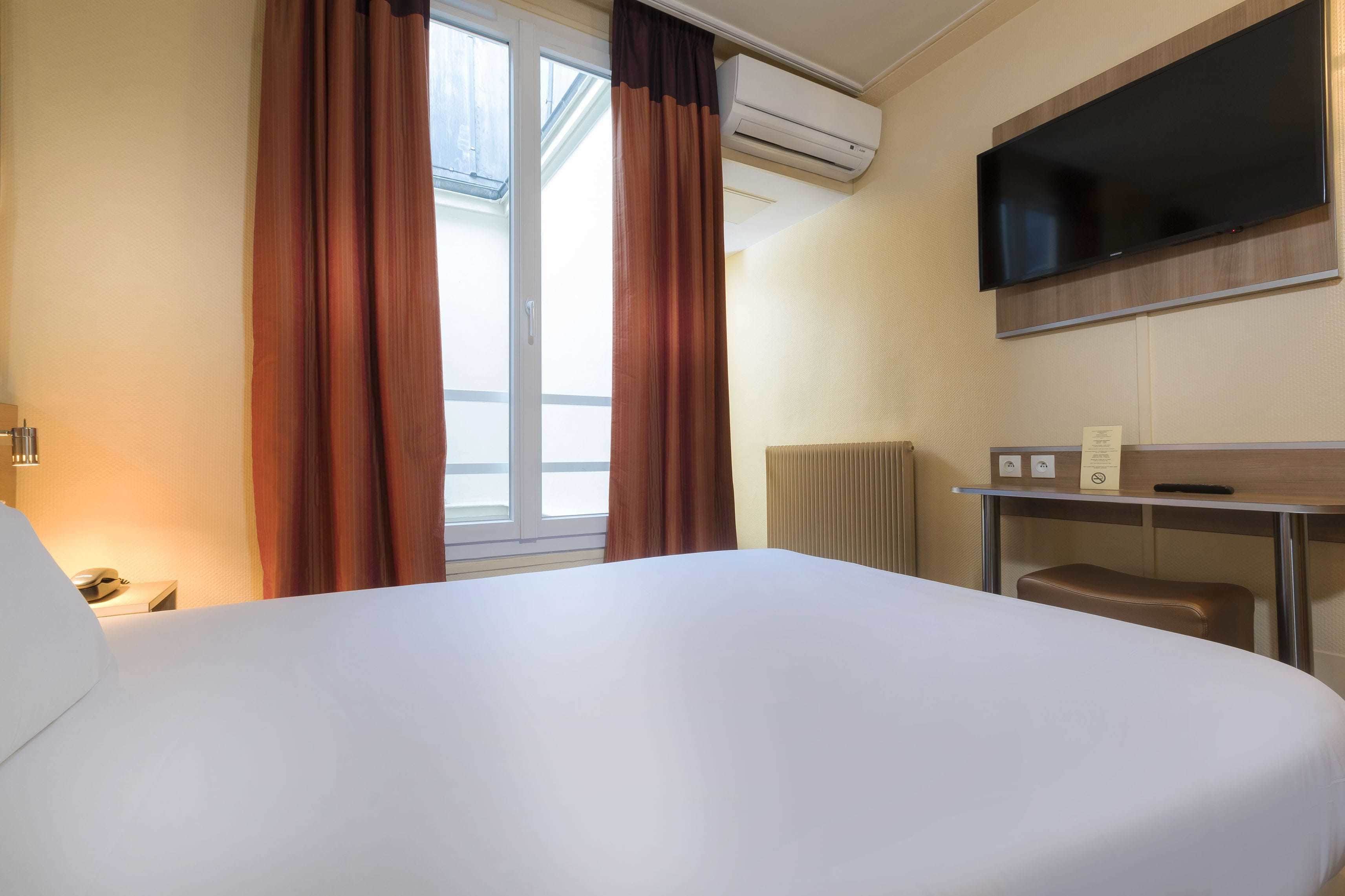 Habitación de hotel con una cama grande, un escritorio con silla, televisor de pantalla plana, aire acondicionado y una ventana con cortinas.