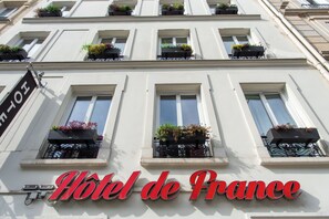 La façade d’un hôtel, ornée de nombreuses fenêtres, dont certaines sont agrémentées de jardinières, et sur laquelle on peut lire en grandes lettres l’inscription « Hôtel de France ».