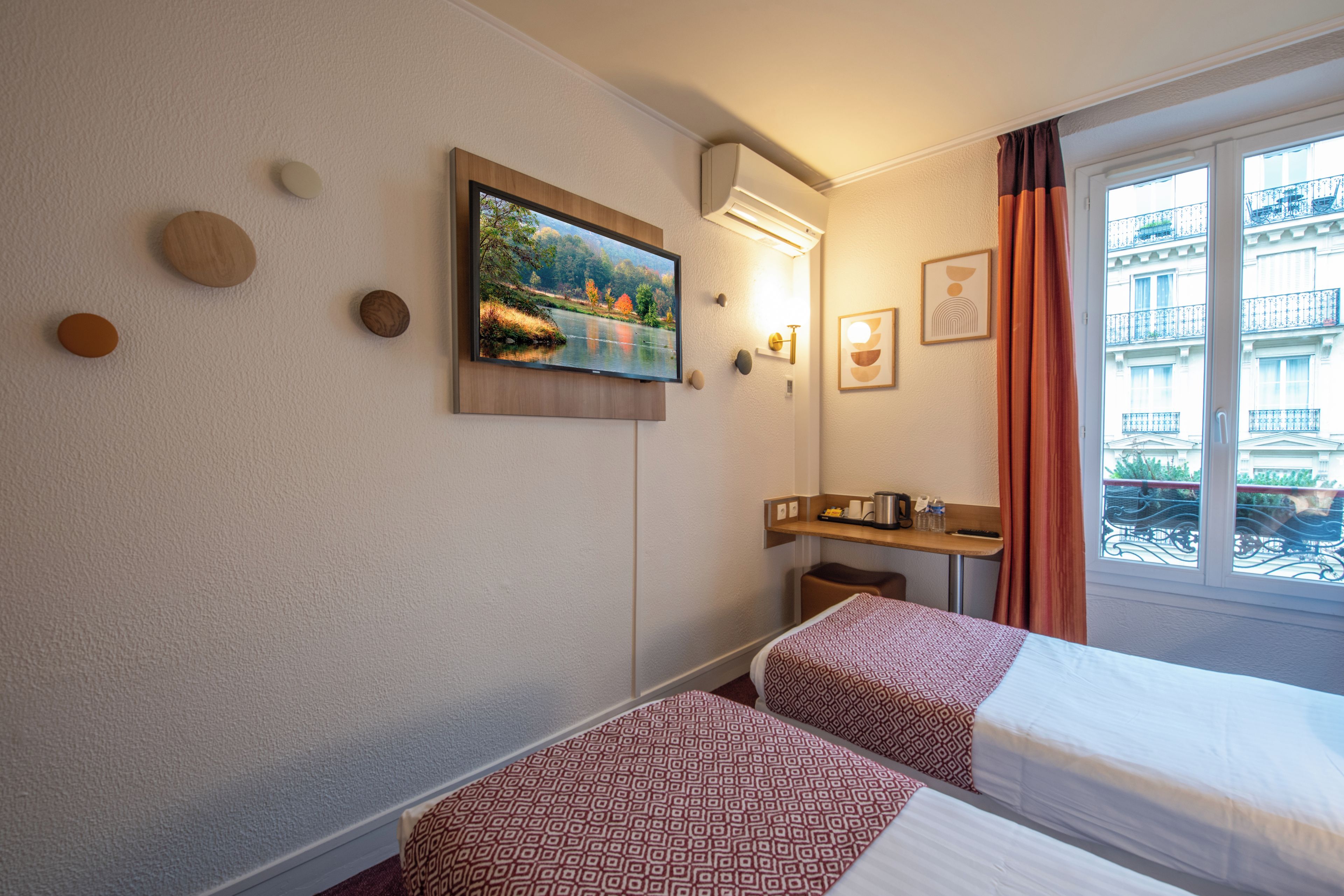 A hotel room with two beds, a painting, a small table with a kettle, and a window with a view of buildings.