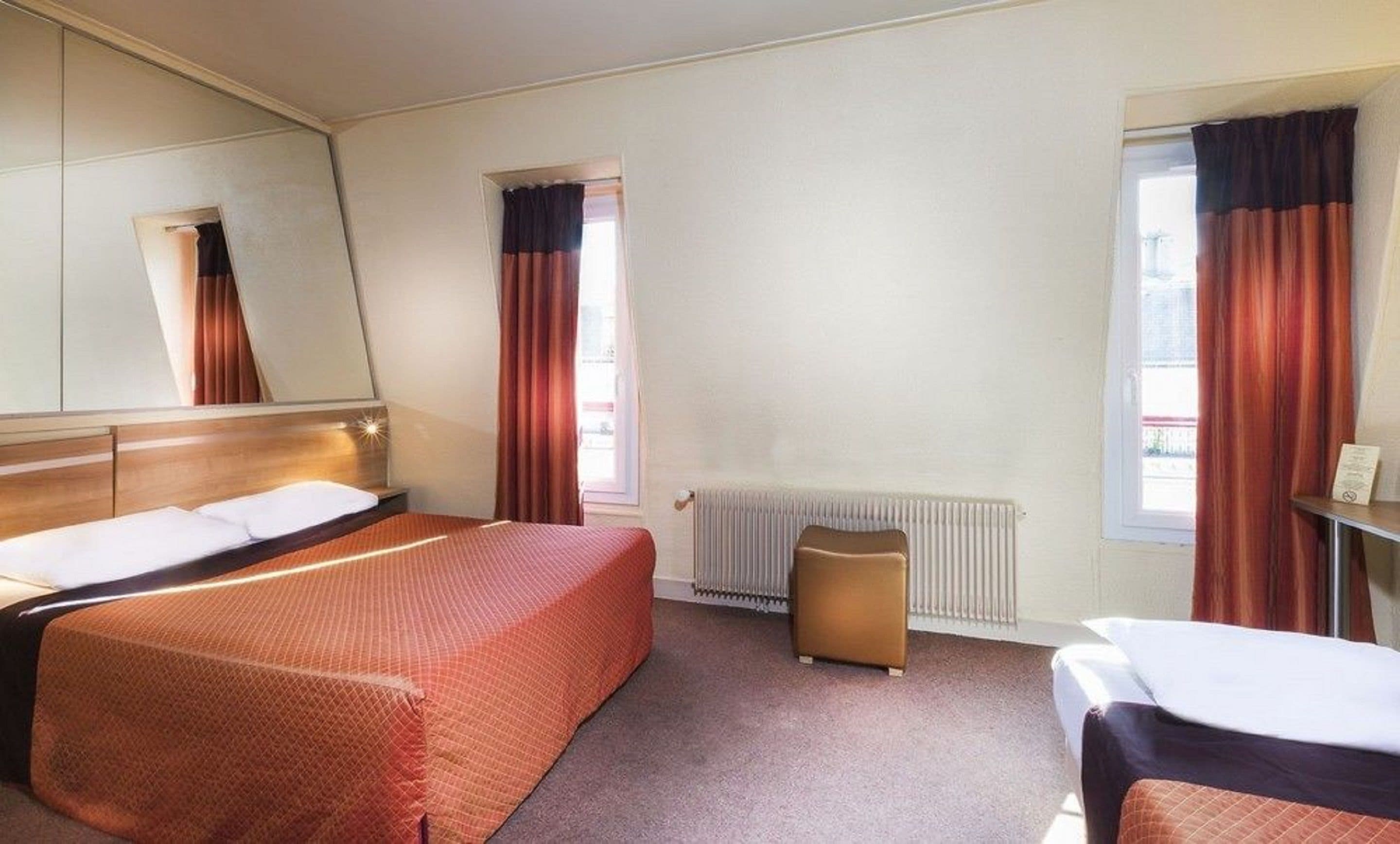 A hotel room with a large bed, a small table, a chair, a radiator, and a window with curtains.