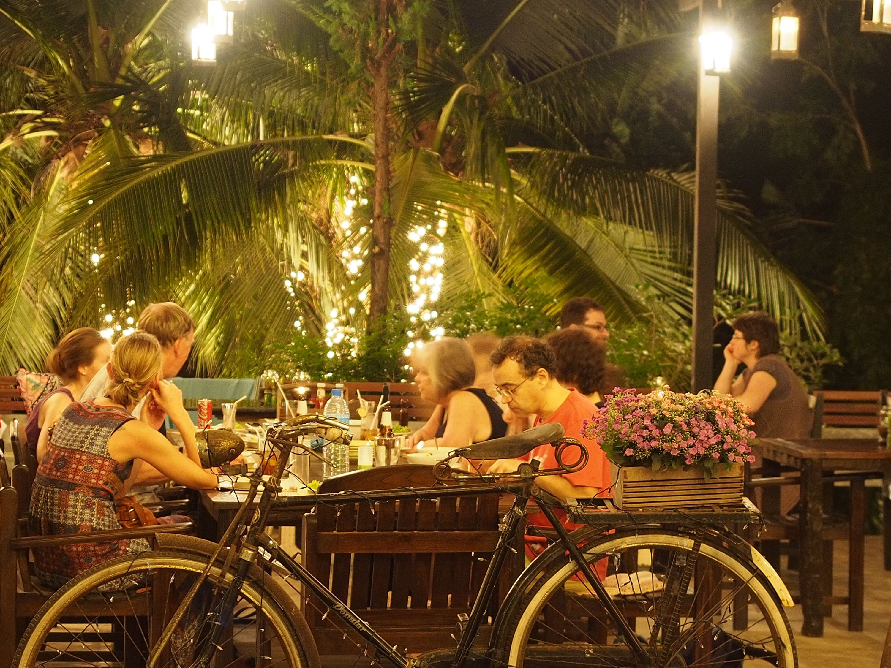 Un grupo de personas cenando al aire libre de noche, con palmeras y luces colgantes al fondo.