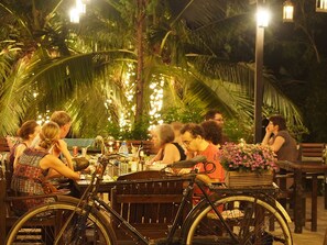 A group of people dining outdoors at night with palm trees and string lights in the background.