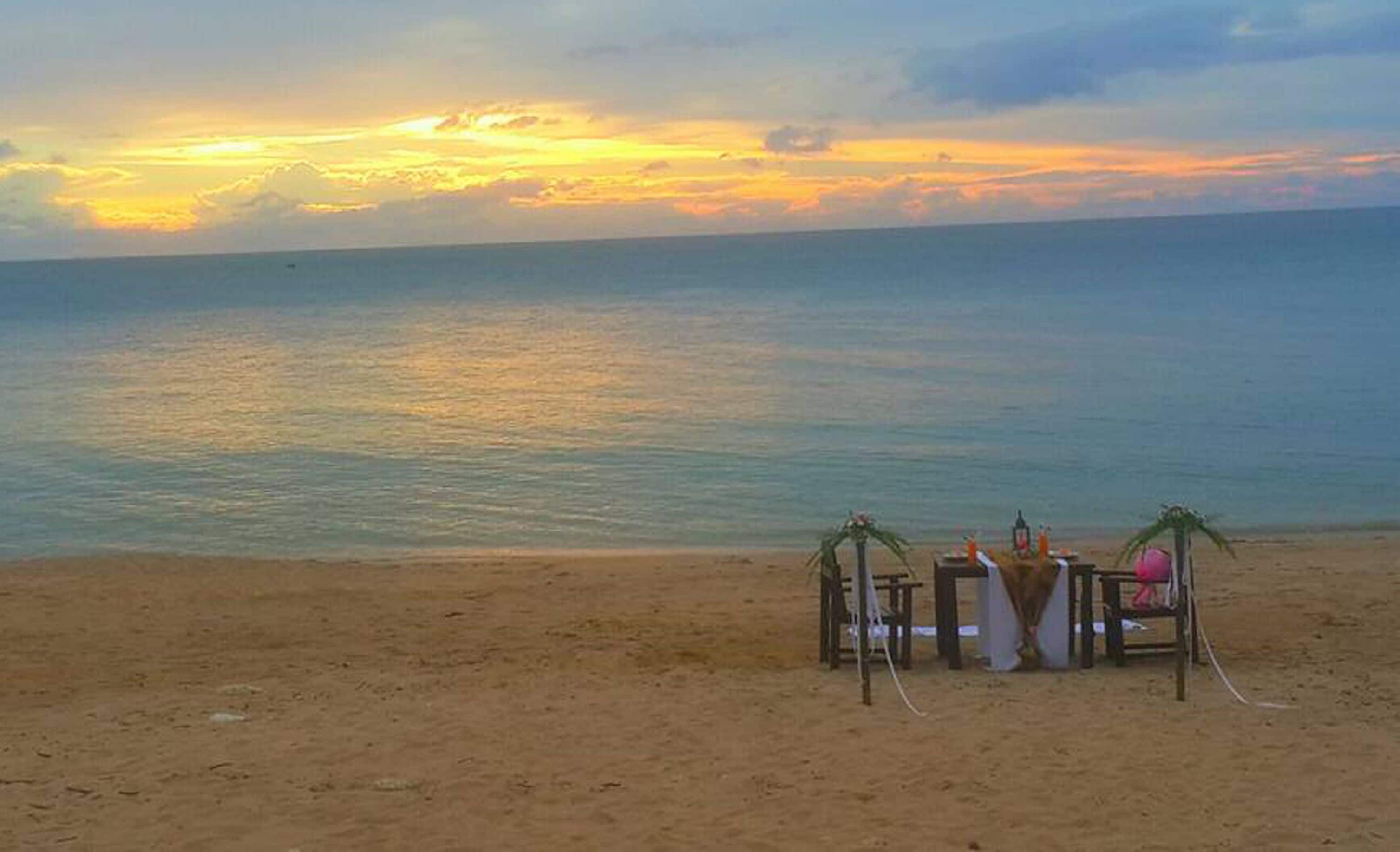 Uma praia com um pôr do sol, uma pequena construção com uma mesa e cadeiras, e uma prancha de surf encostada a ela.