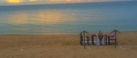 Uma praia com um pôr do sol, uma pequena construção com uma mesa e cadeiras, e uma prancha de surf encostada a ela.