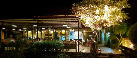An outdoor restaurant area with a covered seating area, a pathway, and a tree with lights.