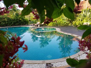 A clear blue pool surrounded by lush greenery, with a wooden deck and lounge chairs.