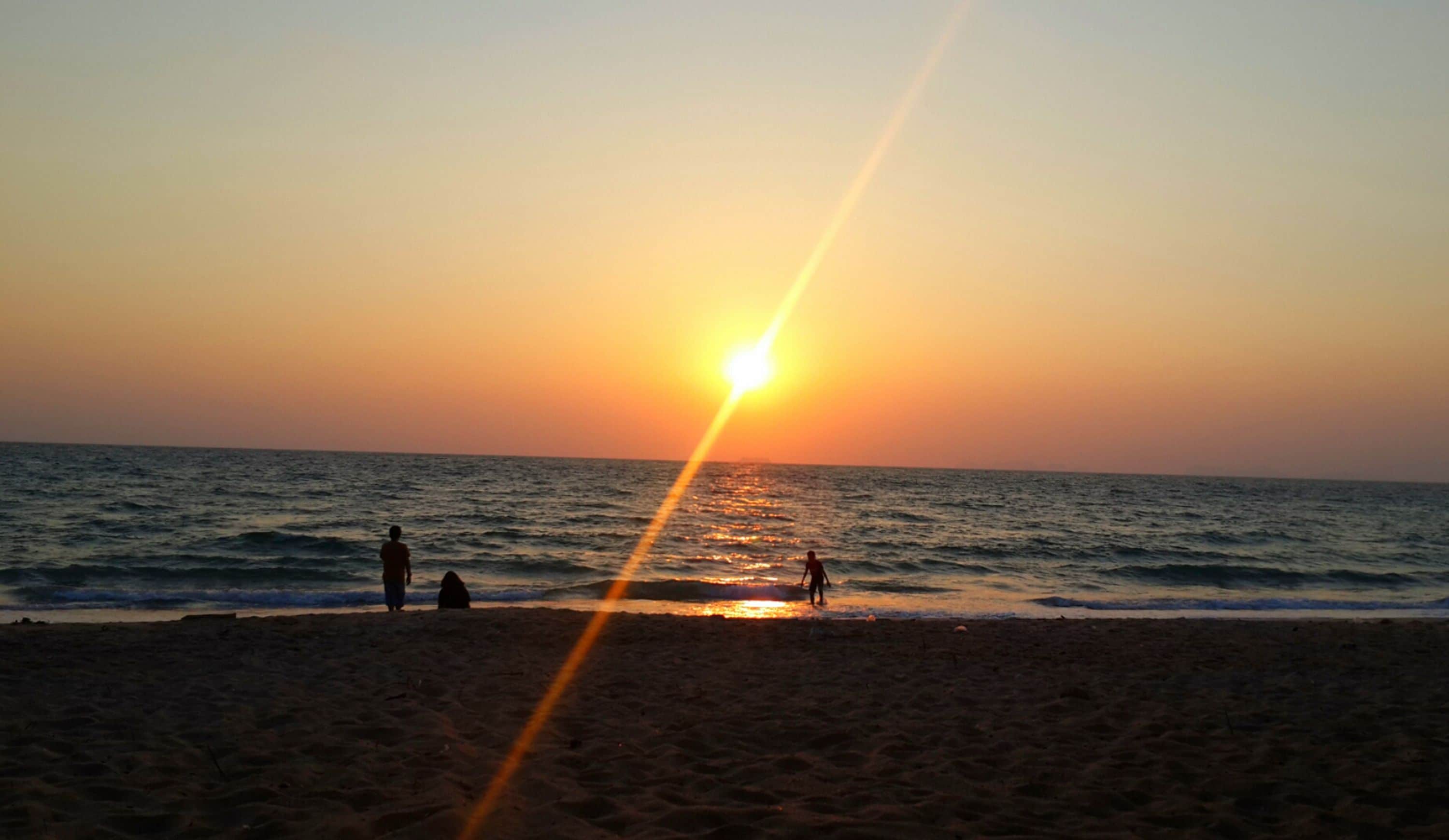 Escena de playa con el sol poniéndose sobre el océano, siluetas de personas en la arena y olas acercándose a la orilla.