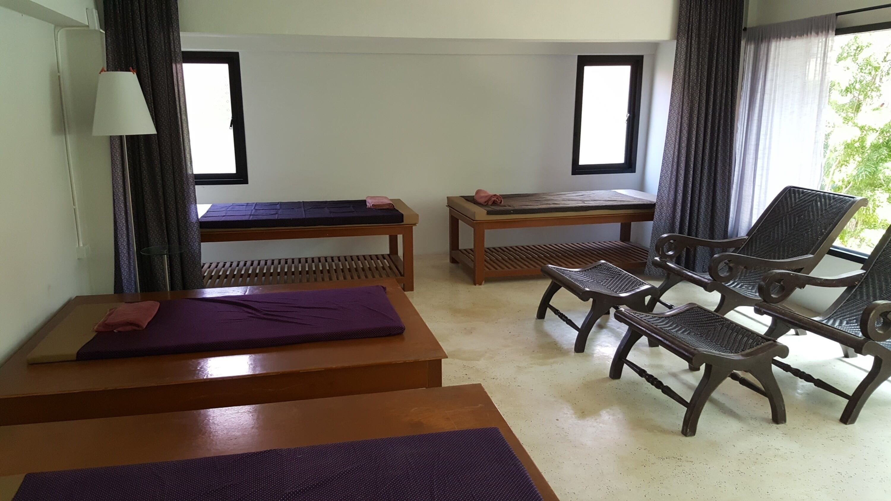 Una habitación con camilla de masaje, sillón de masaje, una mesita con un tapete y una ventana con cortinas.