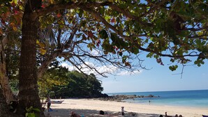 A beach with people sunbathing and swimming, a tree providing shade, and a rocky shoreline in the background. - Tropicana Lanta Resort (Ko Lanta)