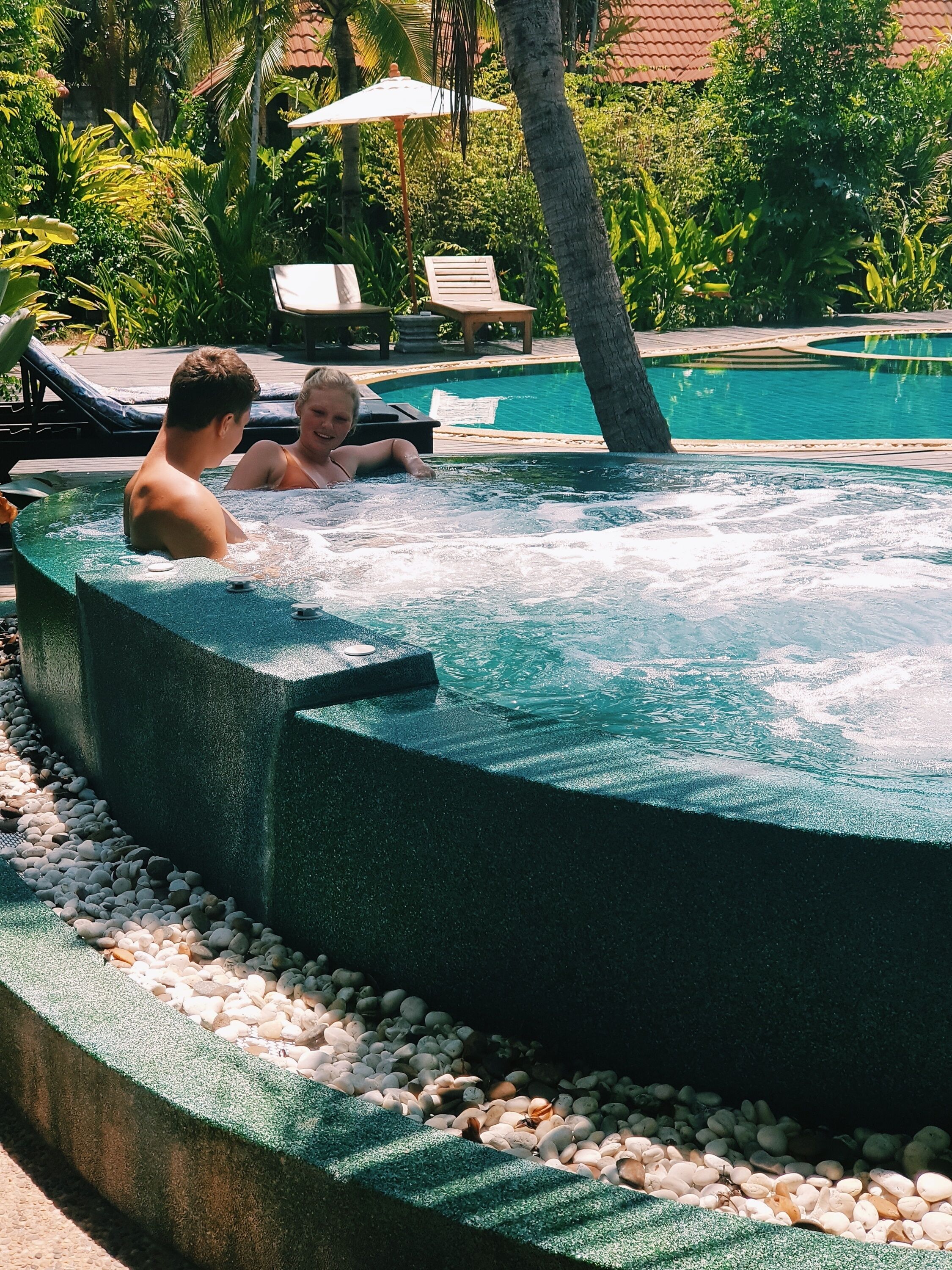 Un bain à remous entouré de galets et de verdure, avec des chaises longues et une piscine en arrière-plan.