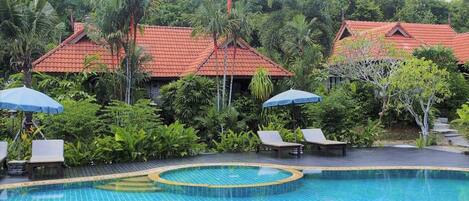 A swimming pool with tiled edges, surrounded by lounge chairs and umbrellas, with red-roofed buildings and lush greenery in the background.
