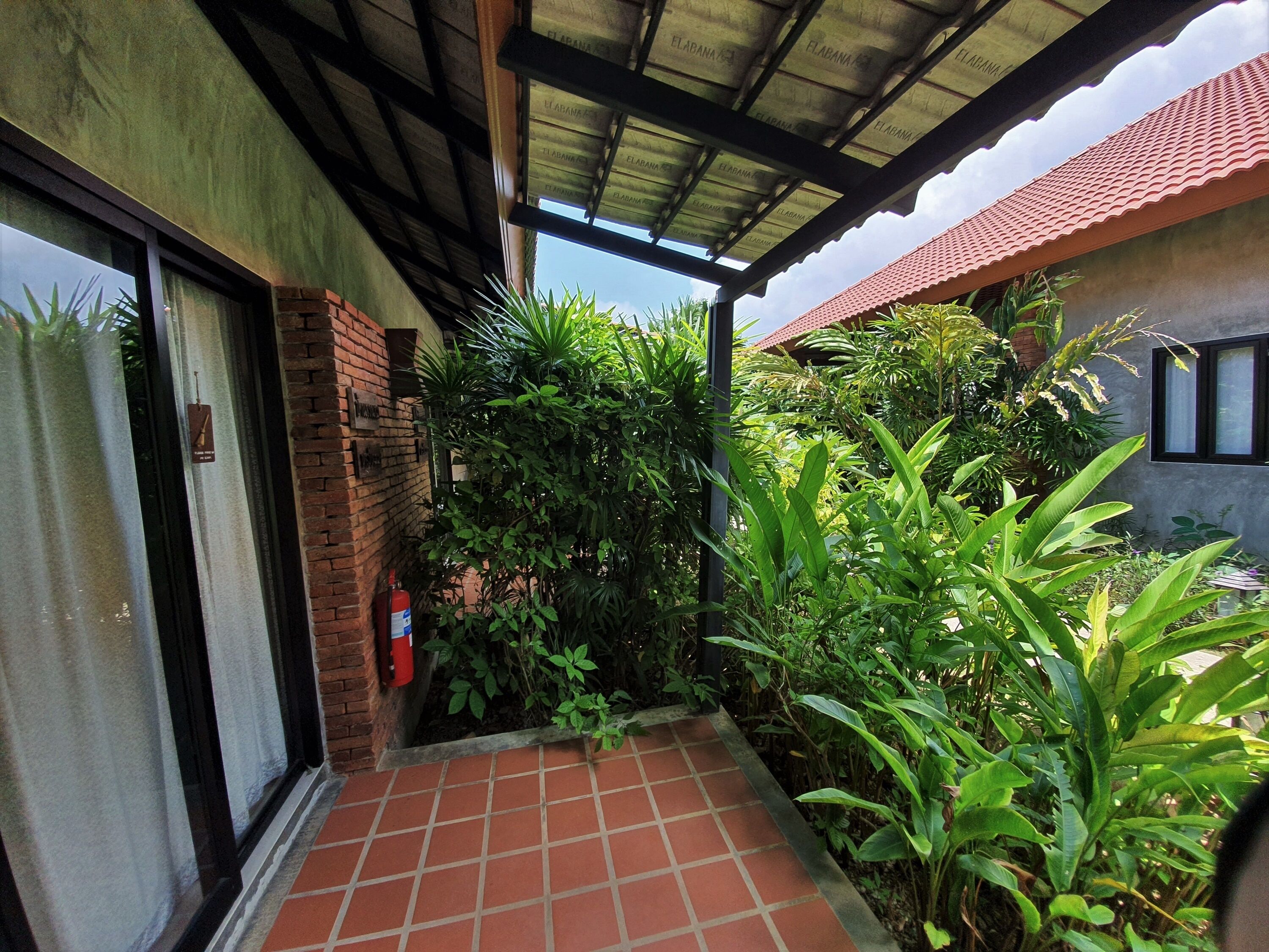 A covered outdoor area with a tiled floor, surrounded by lush greenery and a brick wall.