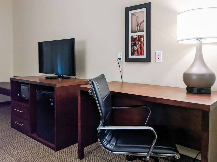 Quarto de hotel com uma escrivaninha de madeira, uma cadeira de escritório preta, uma televisão, um abajur e um quadro na parede.