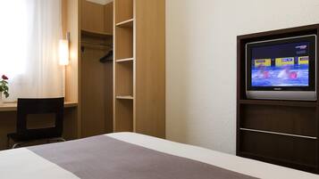 Quarto de hotel com cama, escrivaninha, cadeira, televisão e guarda-roupa.