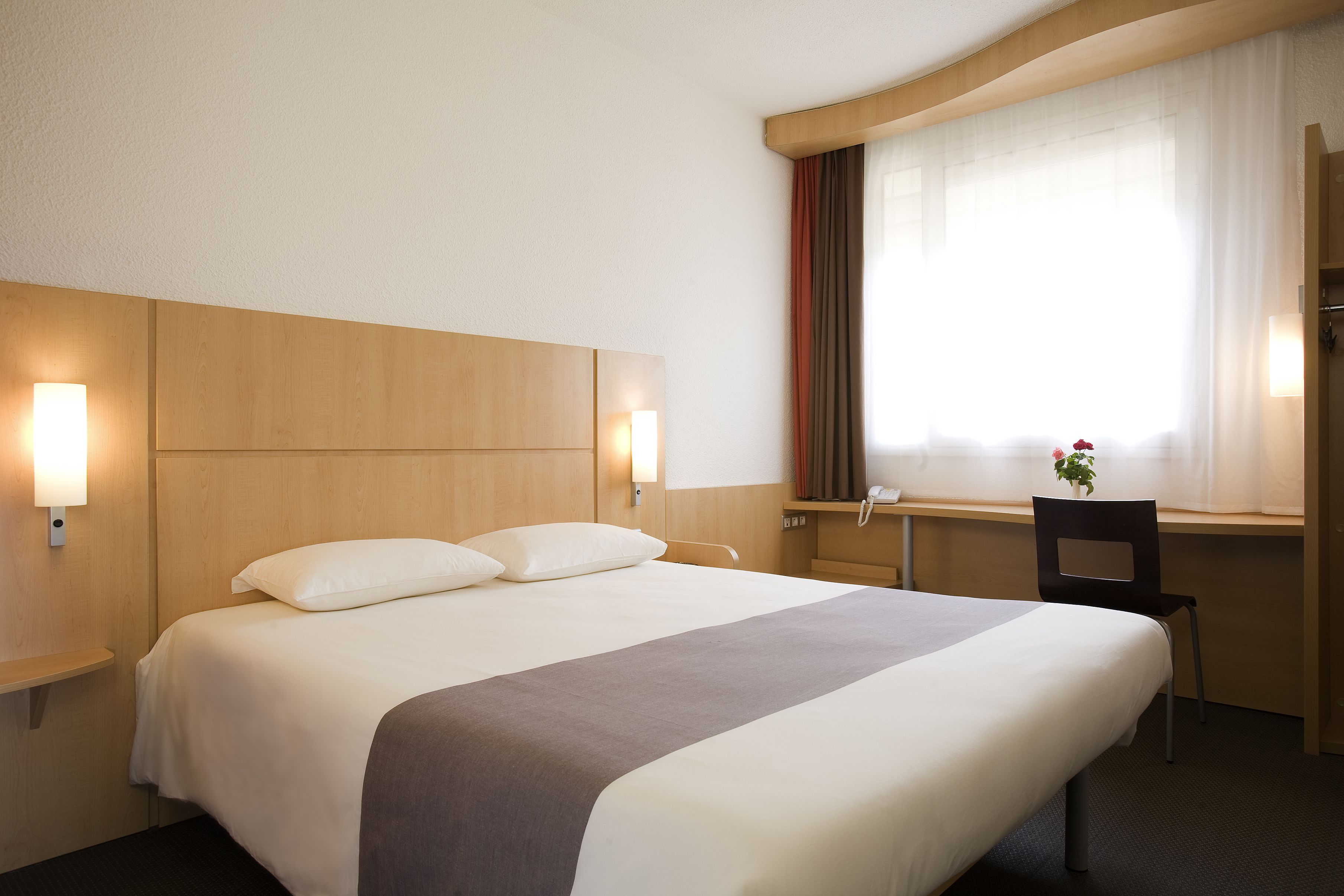 Una habitación de hotel con una cama grande, lámparas de noche, un escritorio con silla y una ventana con cortinas.