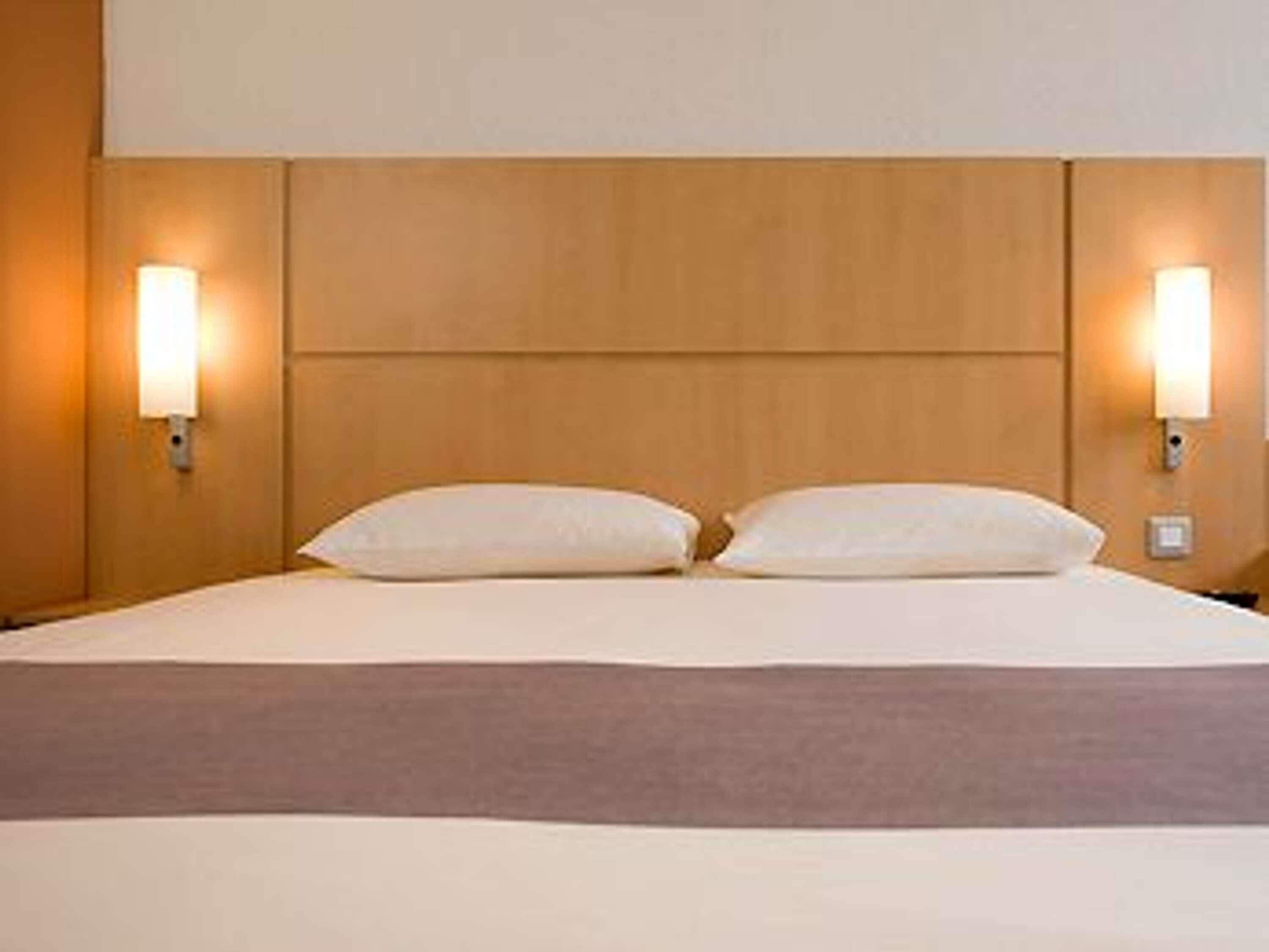 Una habitación de hotel con cabecera de madera, dos lámparas de noche y una cama con ropa blanca.