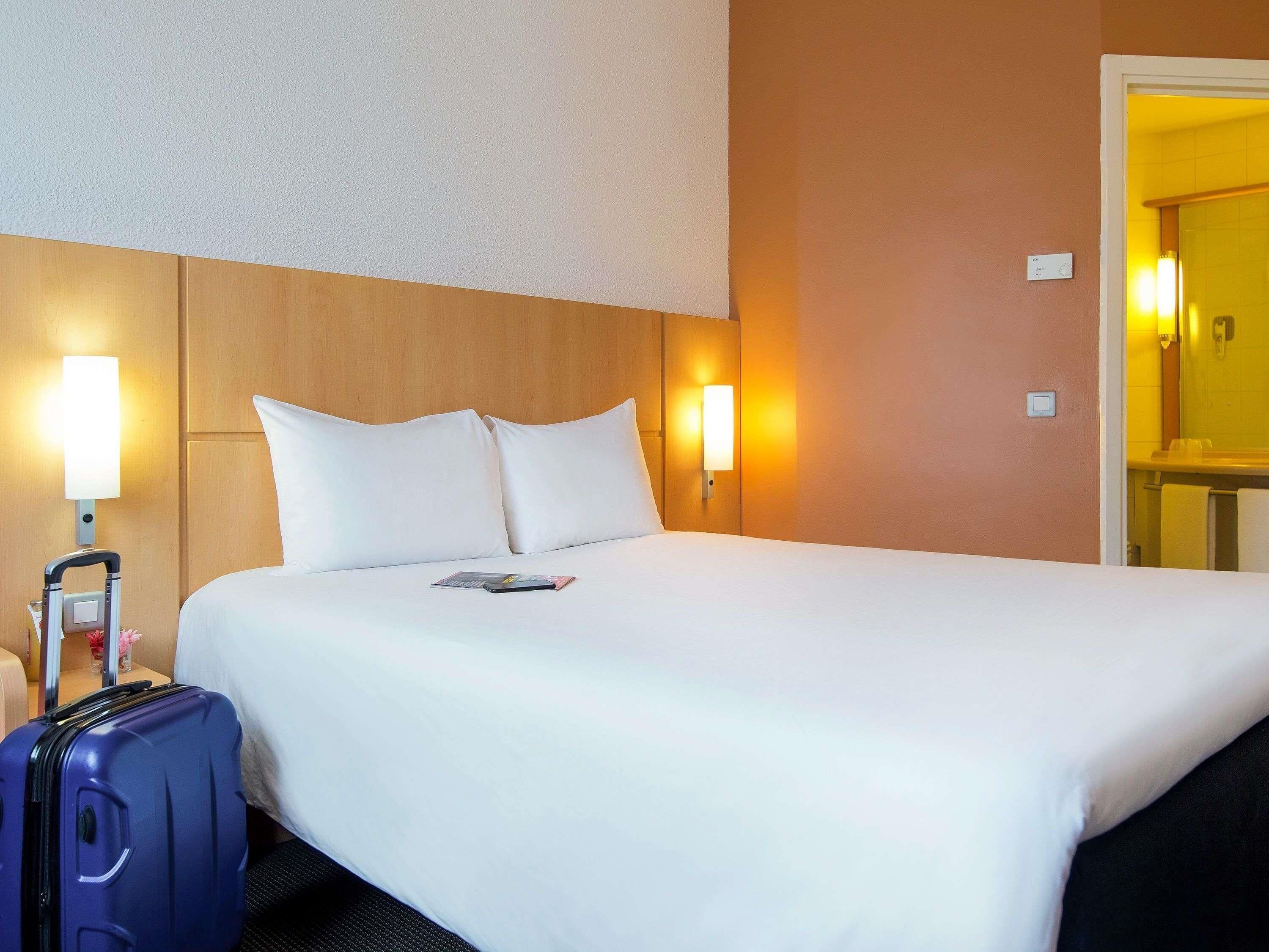 A hotel room with a bed, pillows, a nightstand with a lamp, a suitcase, and a bathroom visible through an open door.
