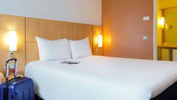 A hotel room with a bed, pillows, a nightstand with a lamp, a suitcase, and a bathroom visible through an open door.