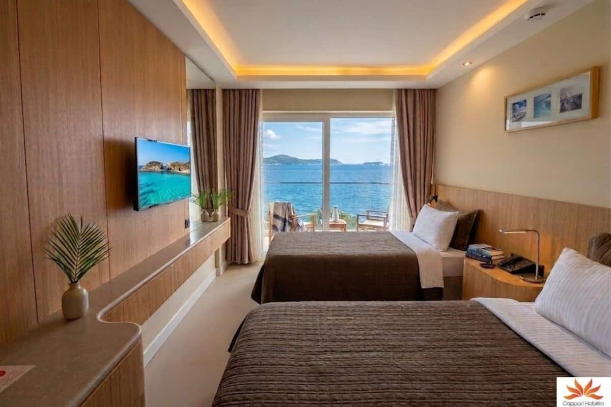 A hotel room with two beds, a TV, a desk, a chair, a window with a view of the sea, and a ceiling light.
