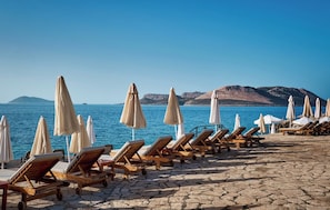 A beachside lounge area with sun loungers and umbrellas, a stone-paved walkway, and a view of the sea and distant mountains.