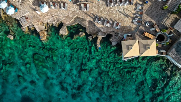An aerial view of a coastal hotel with outdoor seating areas and a clear view of the turquoise sea.
